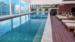 Una piscina interior moderna con agua azul cristalina, un muelle de madera y sillas de descanso.