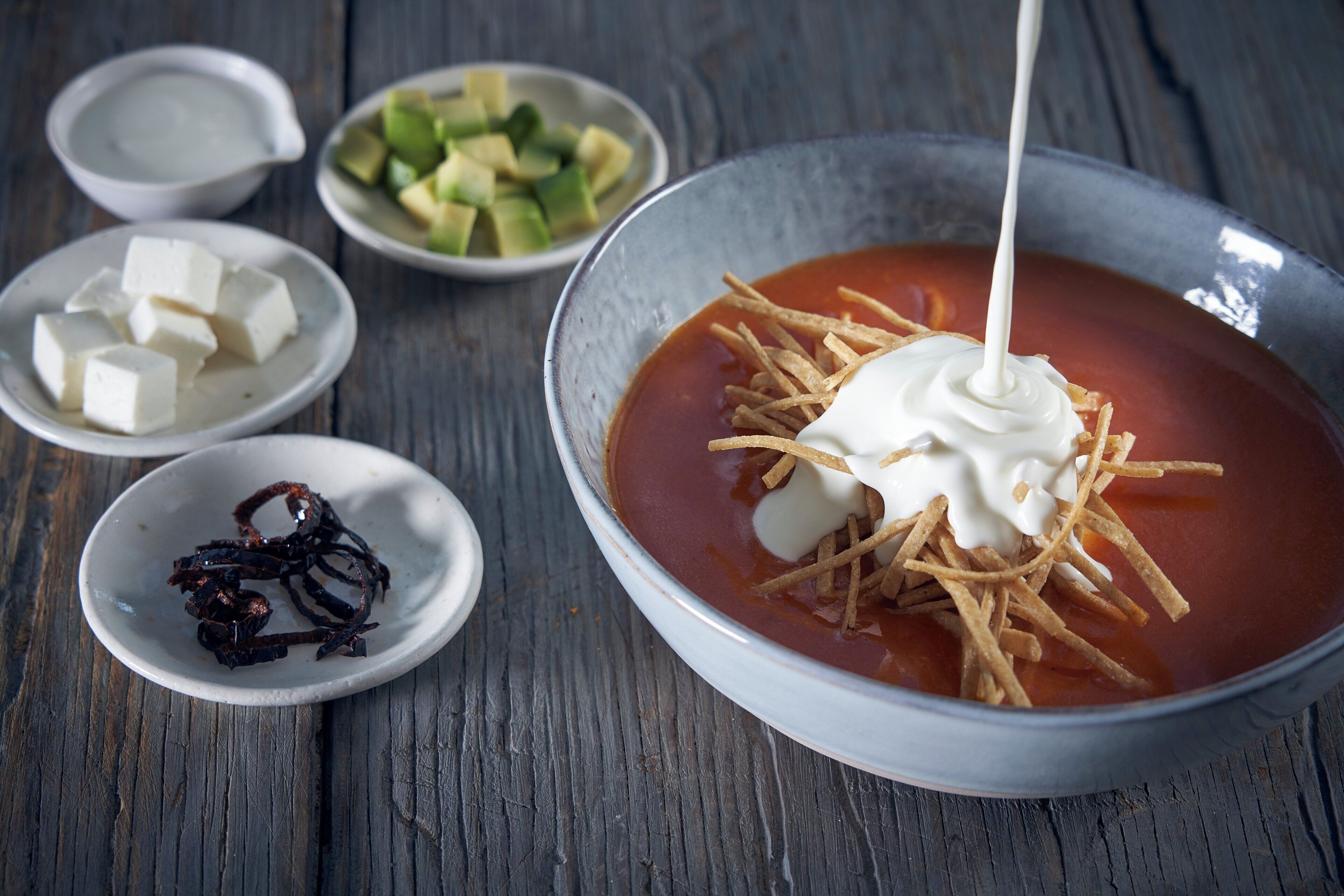 Un tazón de sopa con totopos y crema agria, acompañado de recipientes pequeños con aguacate picado, cubos de queso y camarones secos.