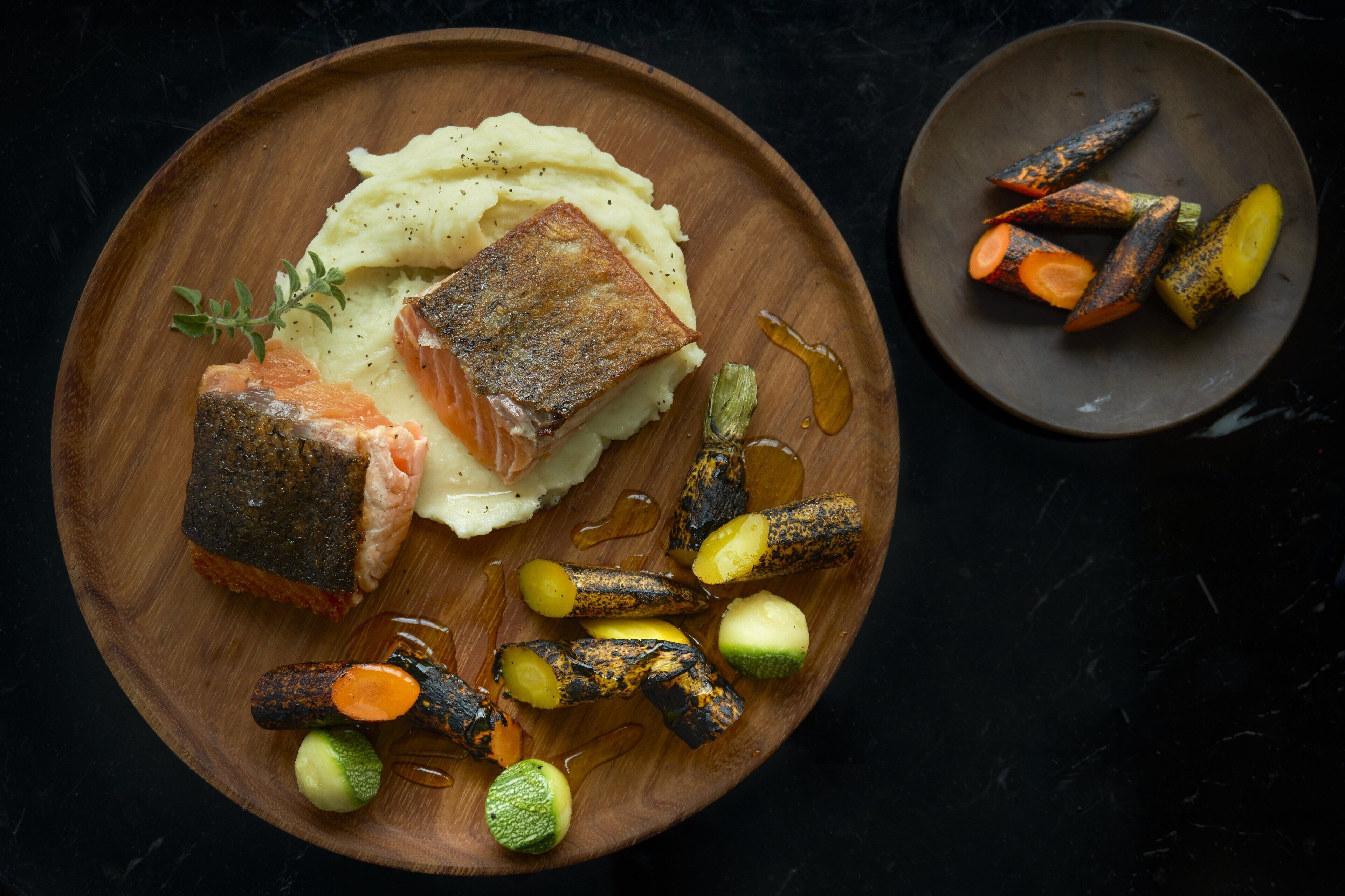 A wooden plate with salmon, mashed potatoes, and roasted vegetables.