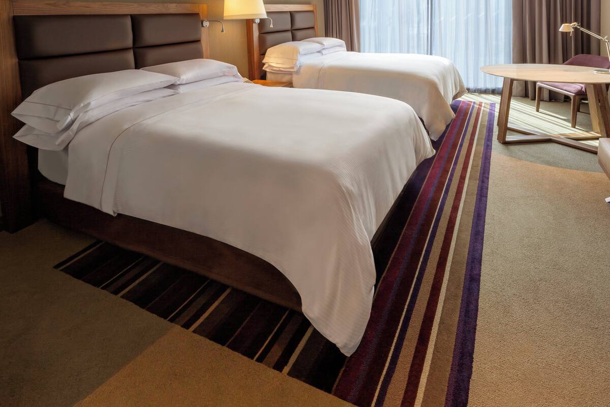 Une chambre d’hôtel avec deux lits, un bureau, une chaise, une lampe et une fenêtre avec des rideaux.