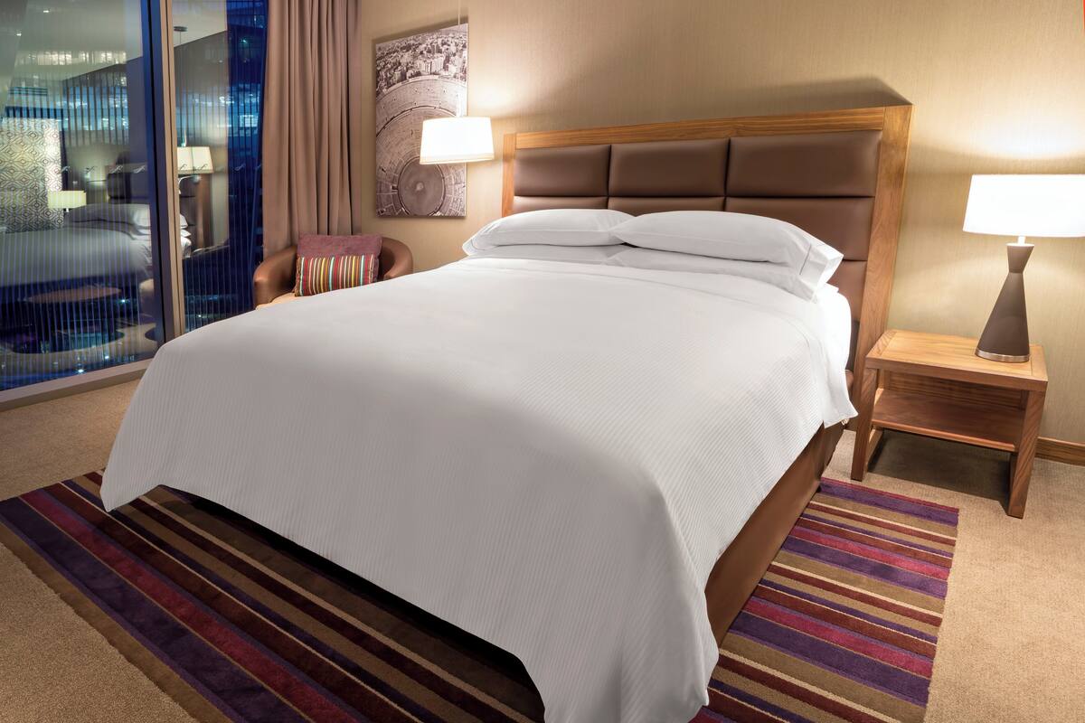 Une chambre d’hôtel avec un grand lit, une table de chevet avec une lampe, un tapis à rayures et une vue sur la ville par la fenêtre.