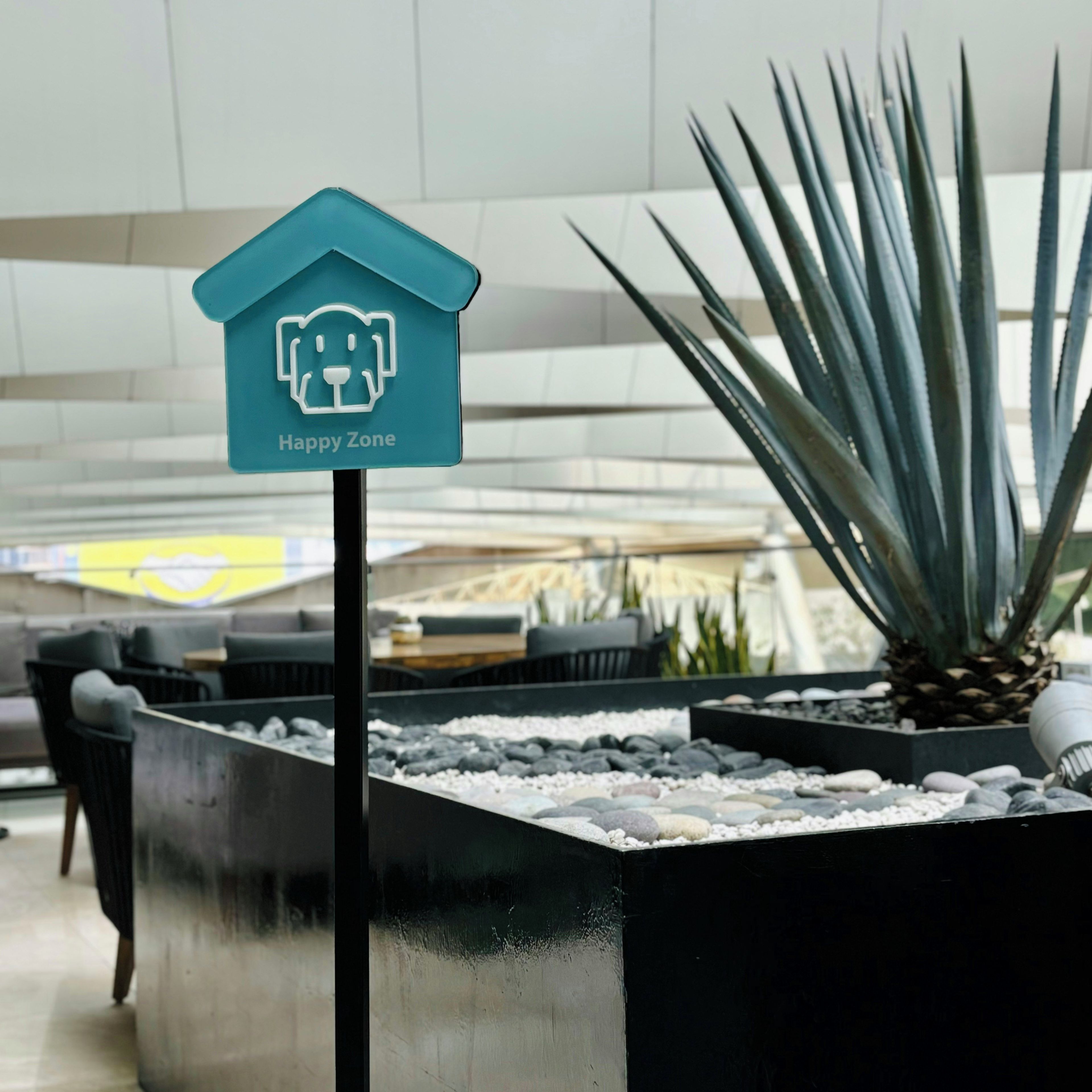 Placa 'Happy Zone' com ícone de urso, planta em vaso e área de estar com piso de pedra.