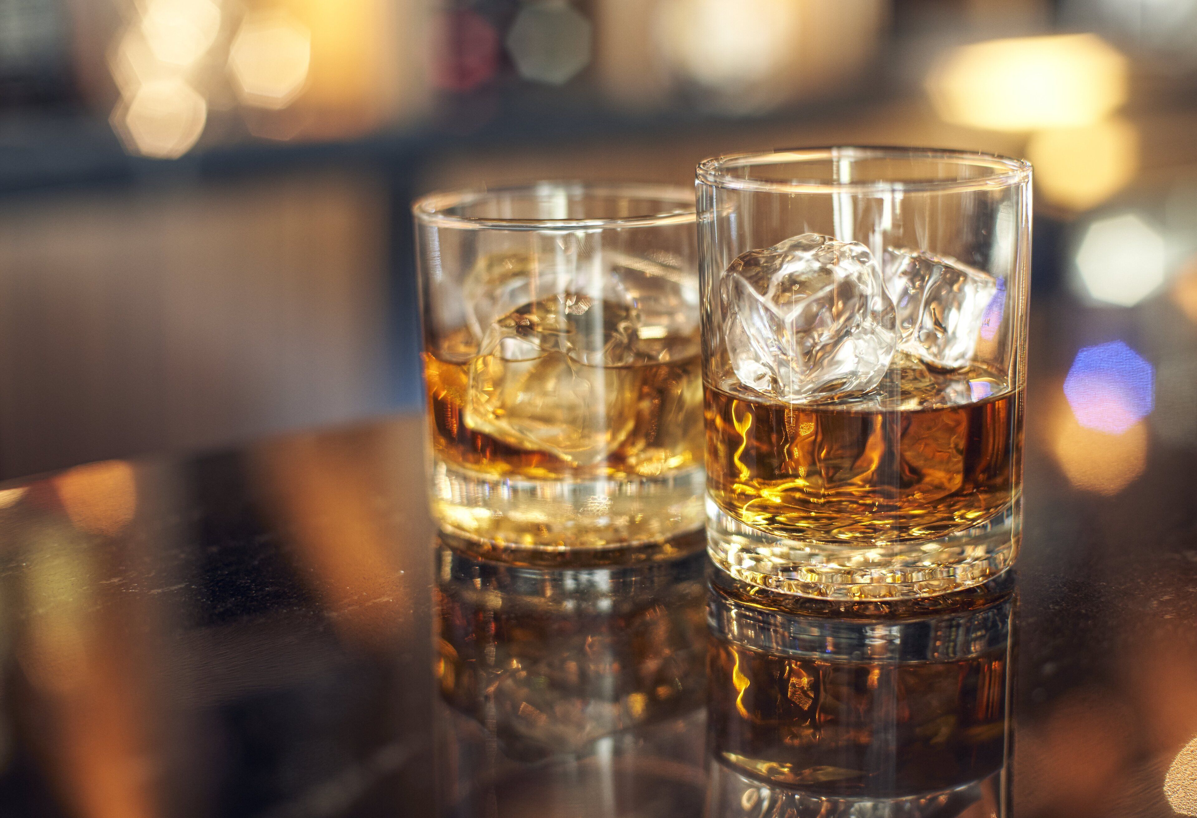 Two glasses of whiskey with ice cubes on a reflective surface.