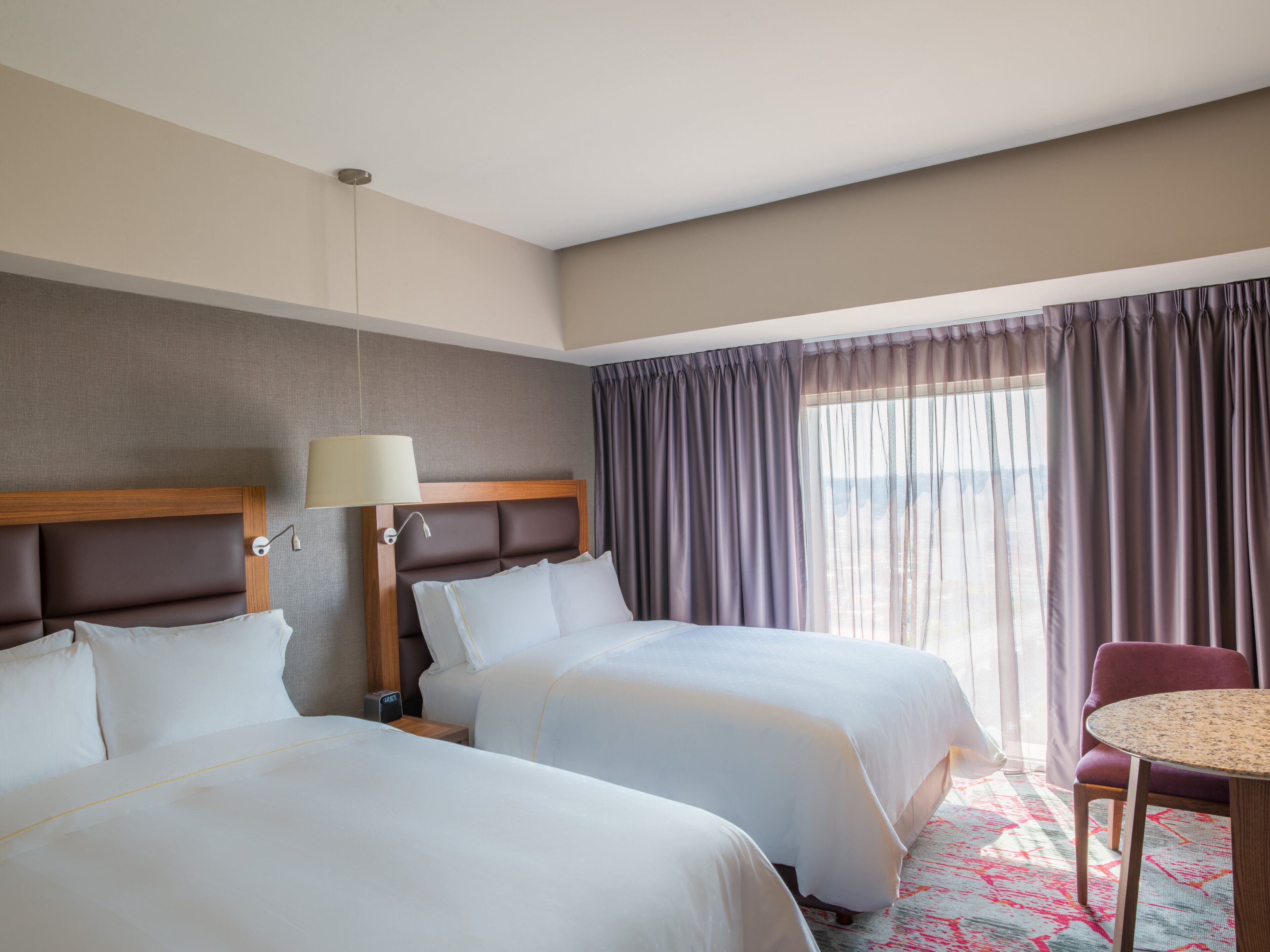 A hotel room with two beds, a nightstand, a lamp, a chair, and a view of the outdoors.