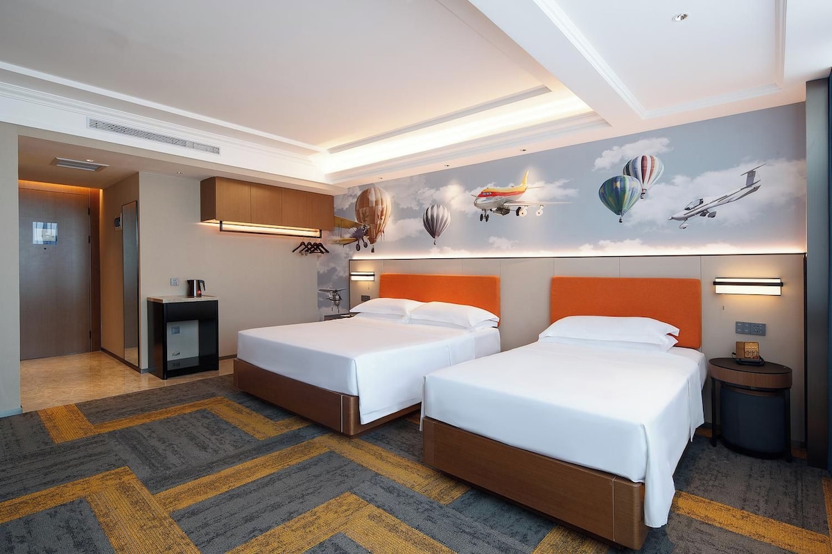 Habitación de hotel con dos camas, una mesita de noche, una lámpara y un mural de aviones en la pared.