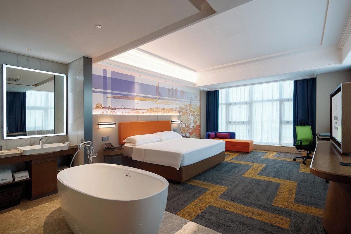 Habitación de hotel con una cama grande, un área de estar, una bañera, un escritorio con computadora y un ventanal amplio.