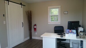A room with a white desk, a black office chair, a fire extinguisher, a window with a view of trees, and a closed white door.
