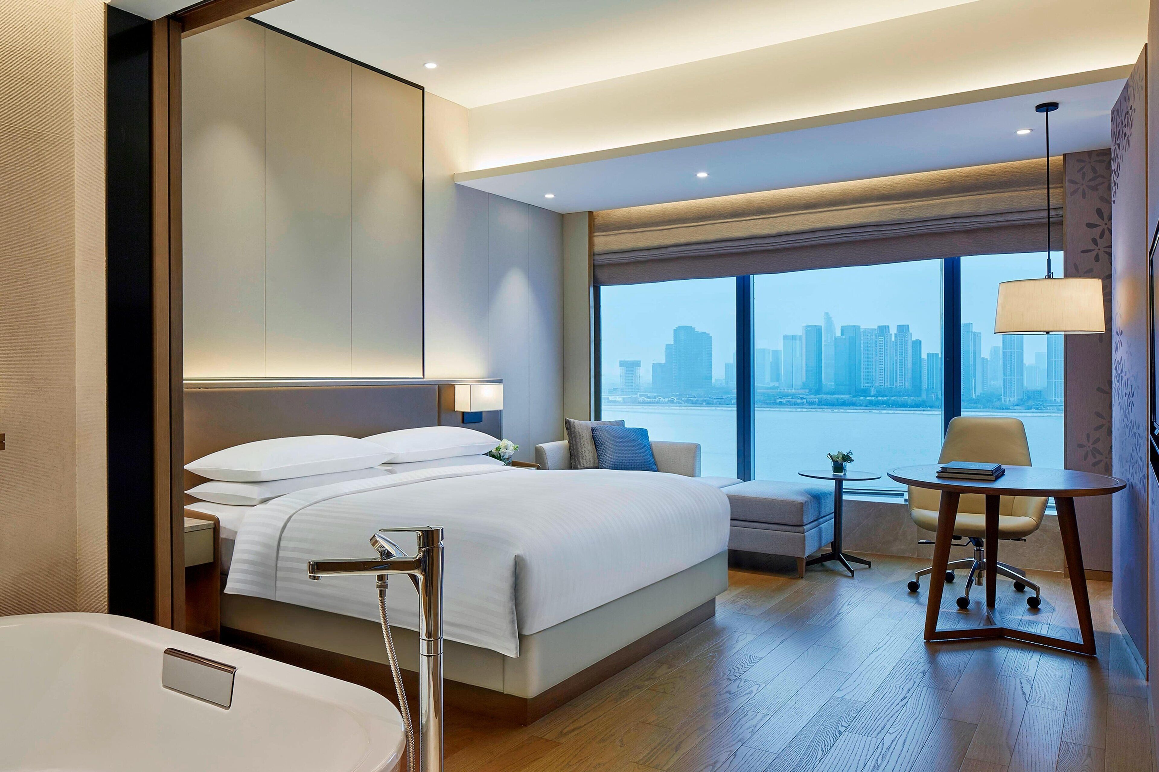 Un hotel moderno con una cama grande, una zona de estar, una mesita y vistas a la silueta urbana.