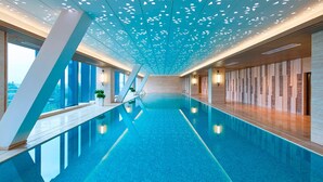 Una piscina interior moderna con diseño geométrico en el techo, ventanas amplias y una pared de madera en un lateral.