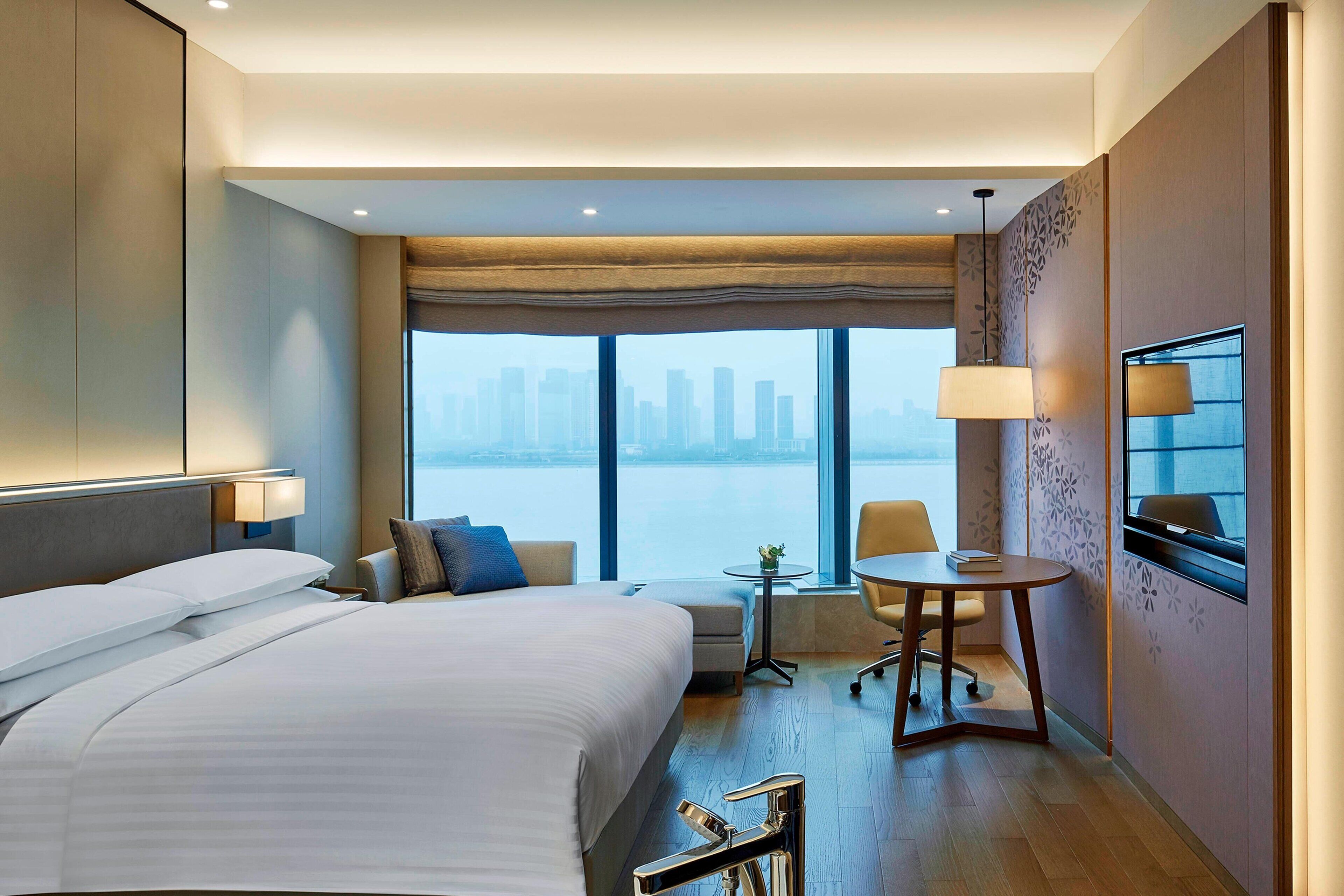 Habitación de hotel con una cama grande, una mesita, una silla y vistas a un horizonte urbano.