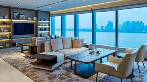 Una sala de estar moderna con un sofá grande, una mesa de centro y un televisor. Hay estanterías con objetos decorativos y un amplio ventanal con vista a la ciudad.