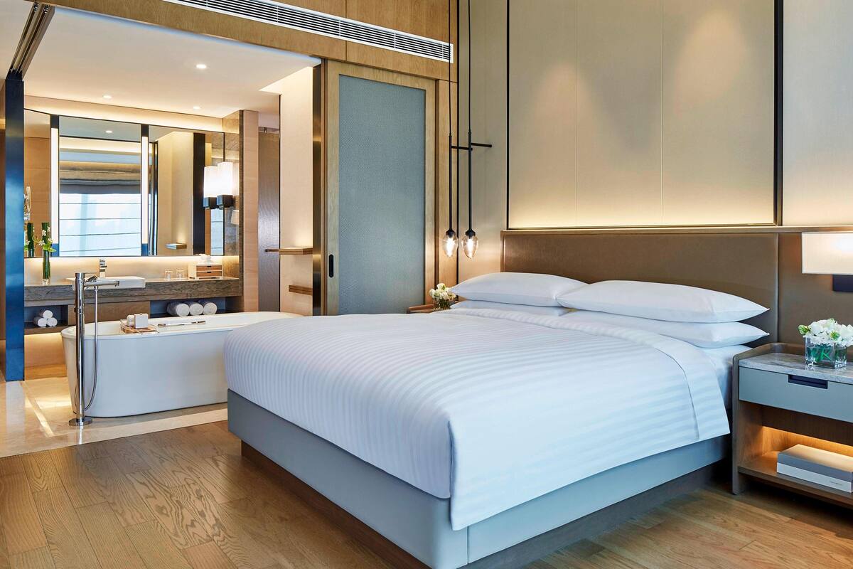 Una habitación de hotel moderna con una cama grande, baño independiente, tocador con espejo y mesita de noche con lámpara.