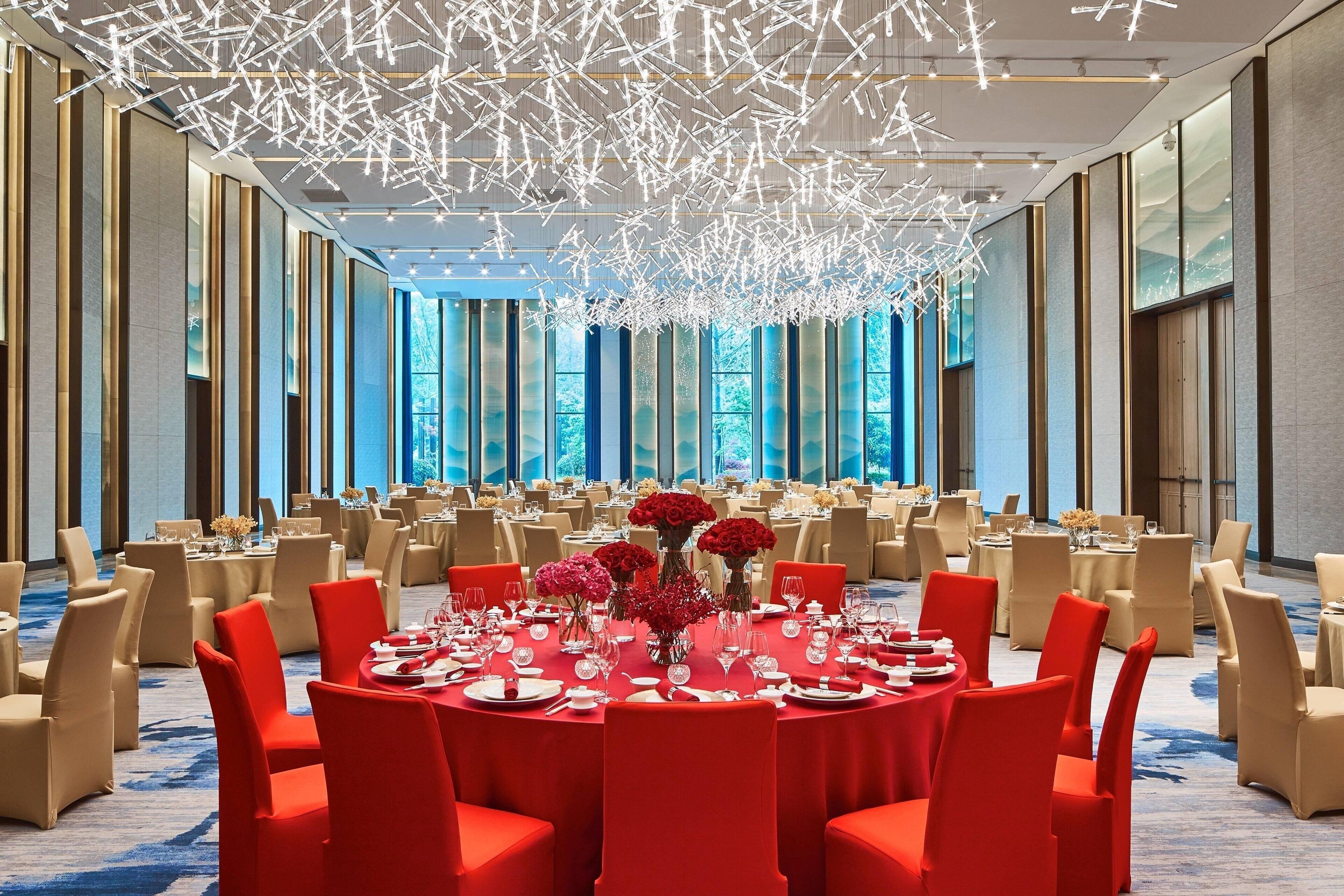 A spacious banquet hall with round tables, red chairs, and a modern chandelier.