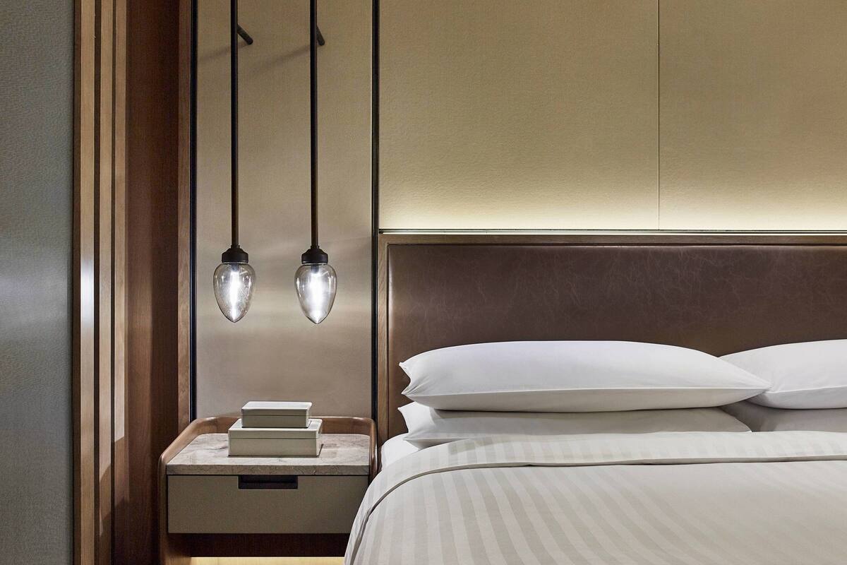 Una cama bien tendida con sábanas blancas y un cabecero, una mesita de noche con libros y una lámpara colgante.