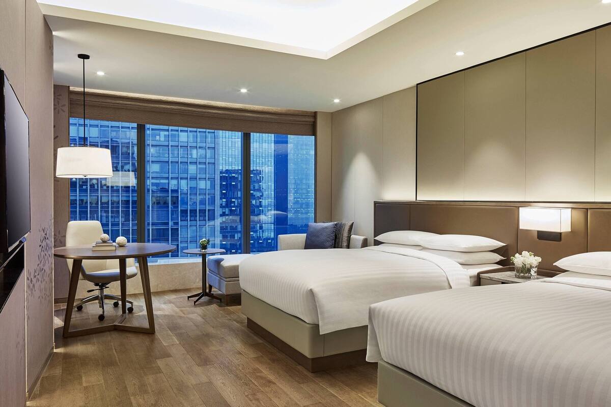 Una habitación de hotel moderna con dos camas, un escritorio y un amplio ventanal con vistas a la ciudad.