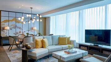 Ein modernes Wohnzimmer mit einem Ecksofa, einem Couchtisch und einem Fernseher. Es gibt einen Essbereich mit Stühlen und einem Tisch, sowie ein großes Fenster mit Vorhängen.