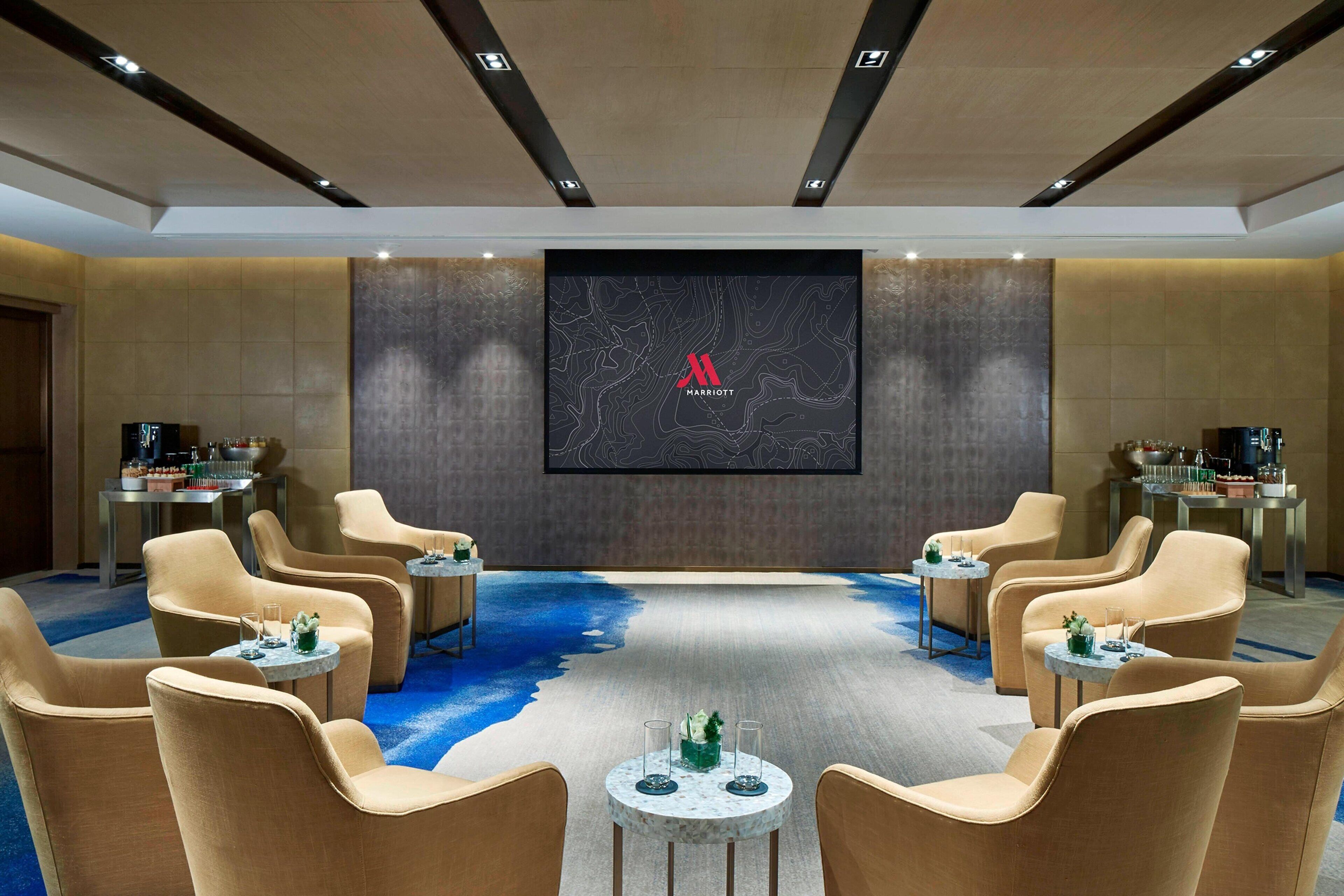 Een vergaderzaal met een groot scherm waarop het Marriott logo staat, beige stoelen rond een centrale tafel gerangschikt, en een blauw tapijt met een waterachtig patroon.