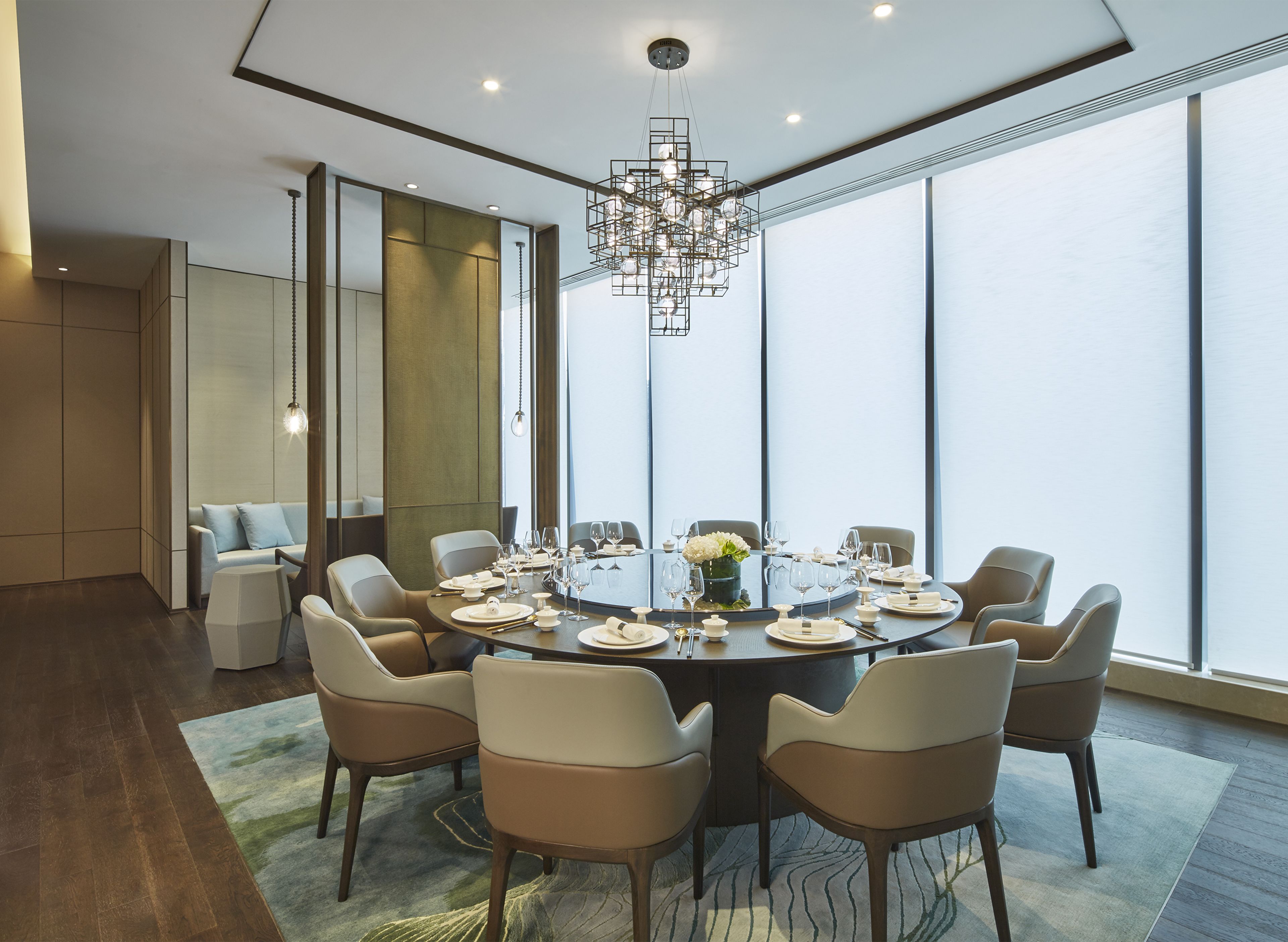 Uma sala de jantar moderna com uma mesa redonda posta para oito pessoas, rodeada por cadeiras, num espaço com grandes janelas e um candelabro.