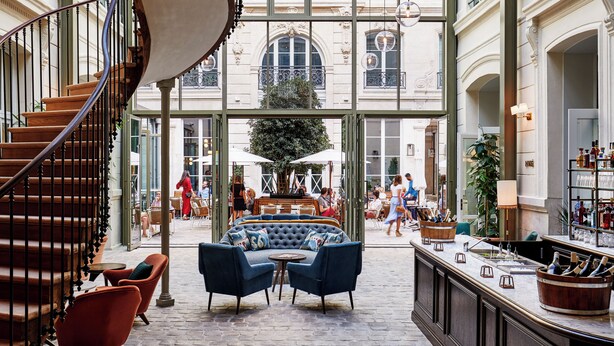 A spacious indoor area with a curved staircase, a seating area with blue sofas, and a bar with bottles. - The Hoxton, Paris (Paris)