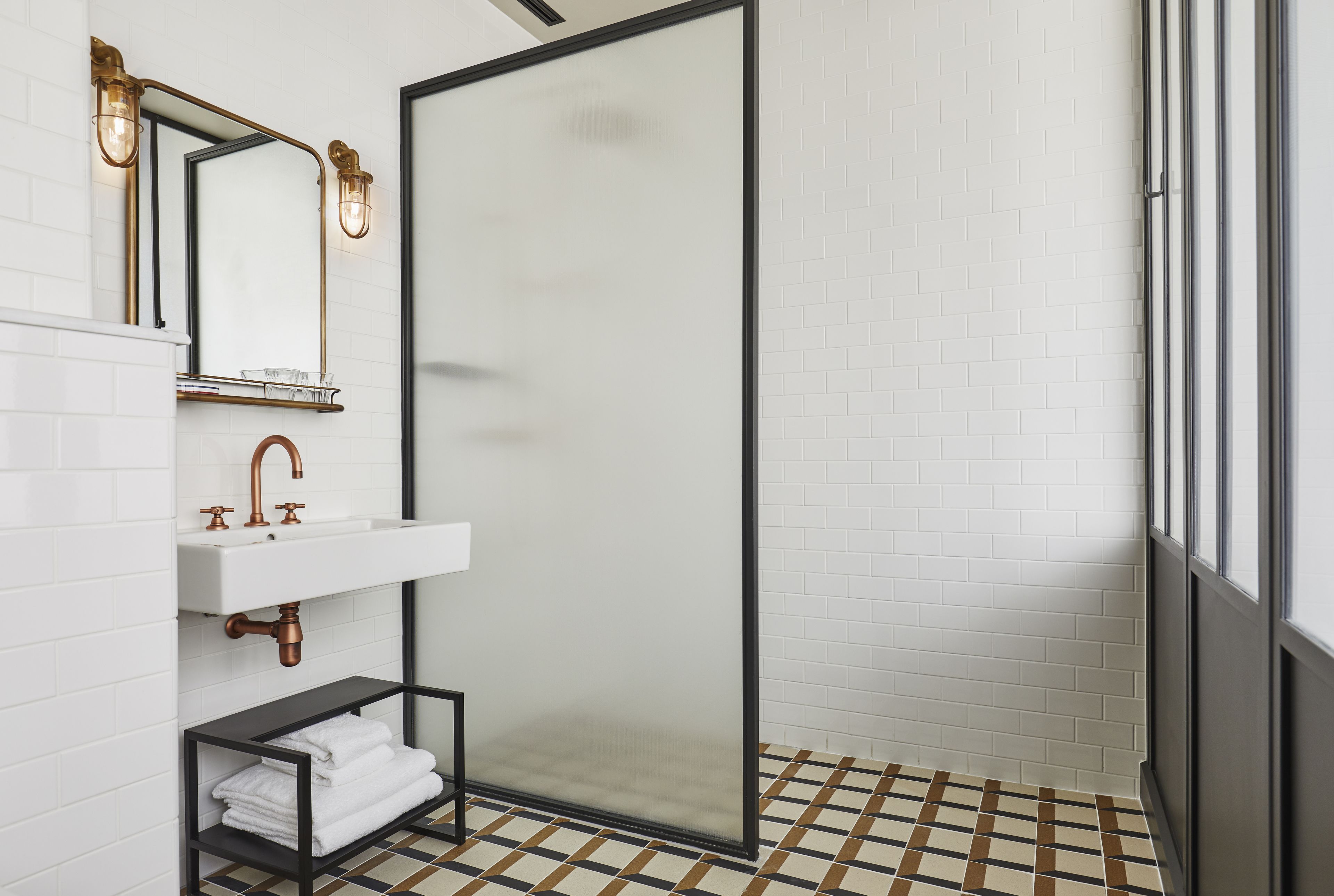 Un bany modern amb sòl de rajoles blanques, un espai de banyera amb portes de vidre, un lavabo blanc amb detalls de coure i una prestatgeria de metall negre amb tovalloles arremolinades.