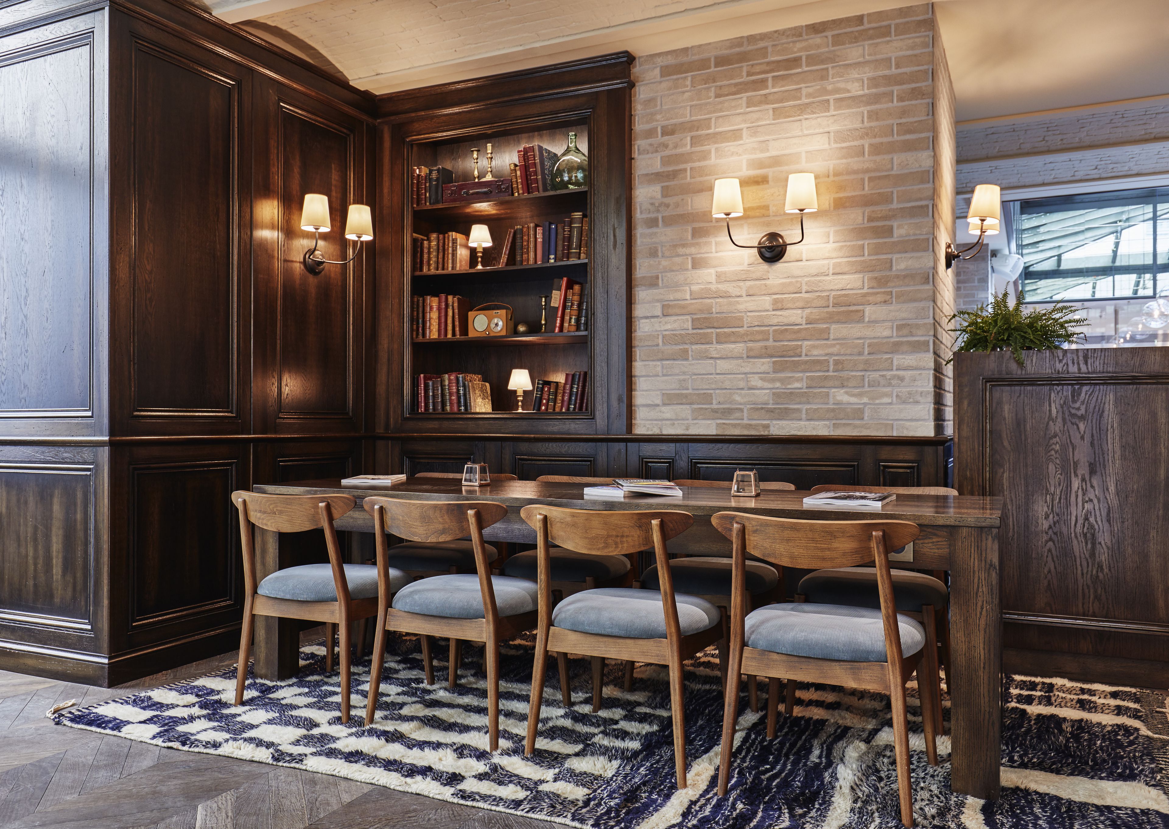A dining area with a wooden table and chairs, a bookshelf, and wall-mounted lighting.