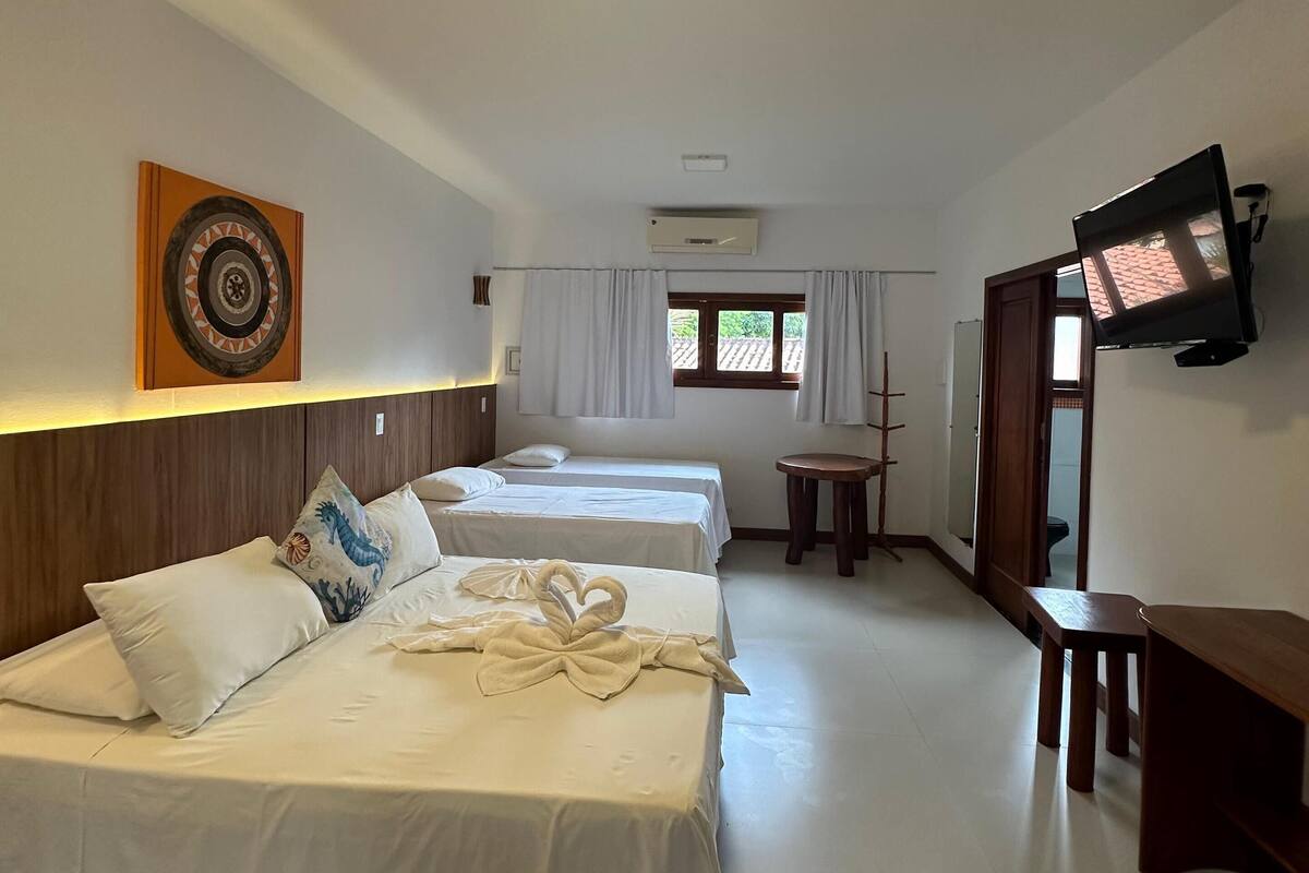 A hotel room with three beds, a flat-screen TV, an air conditioning unit, and a window with curtains.