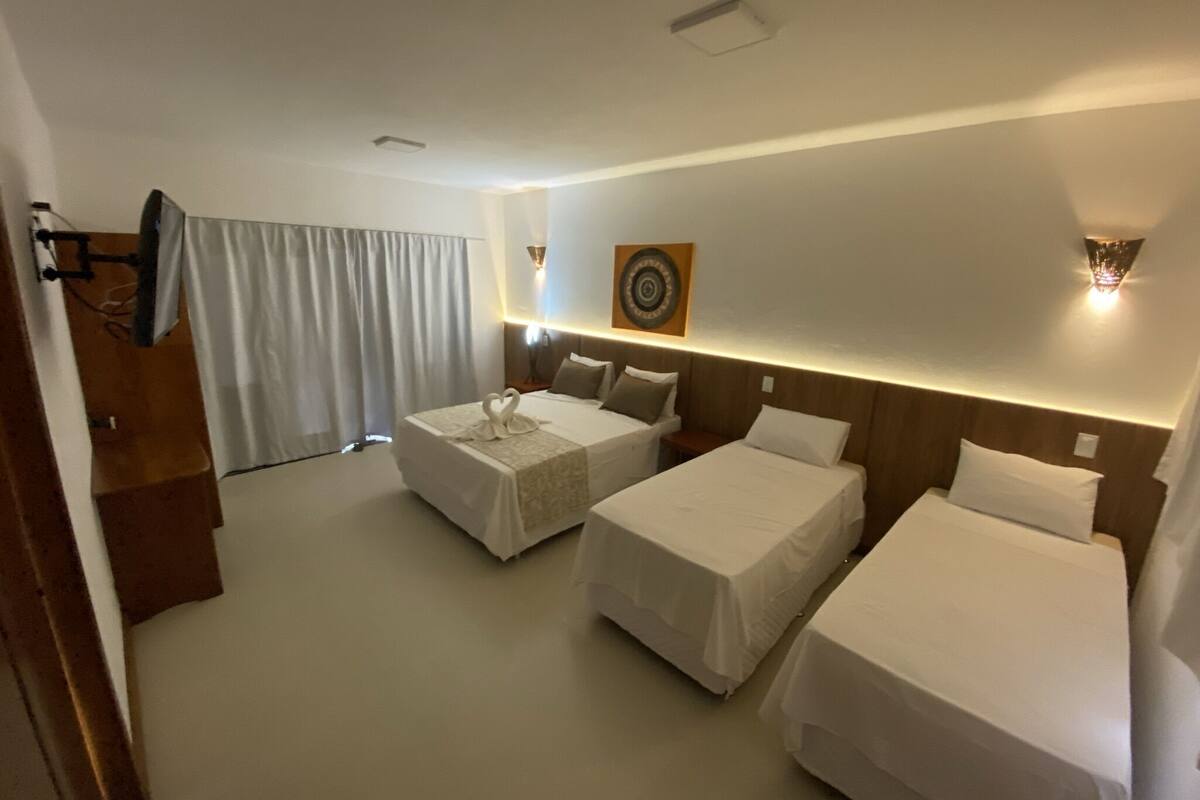 A hotel room with two single beds, a large window with curtains, a wall-mounted clock, and a small table with a lamp.