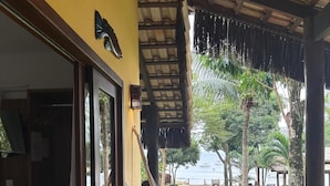 Un porche de madera con una hamaca, una mesa de madera y vistas a una playa y palmeras.