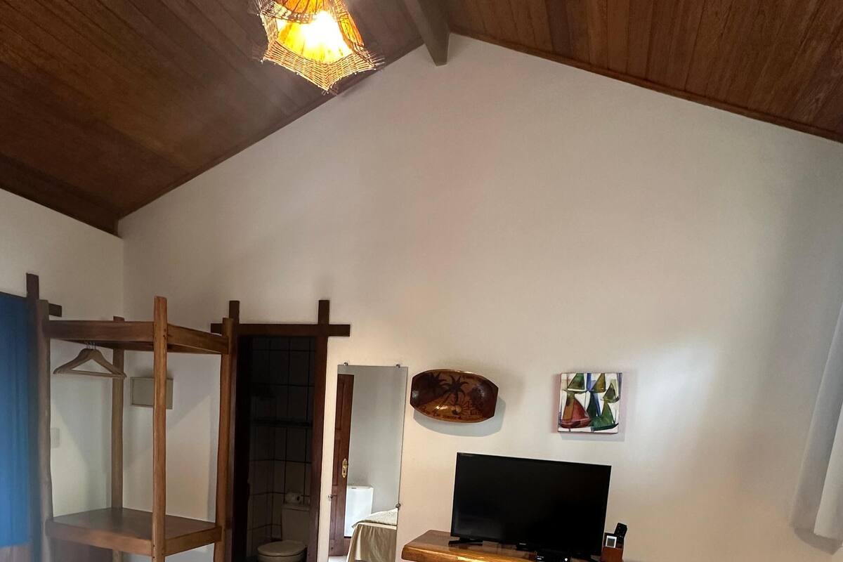A bedroom with a bed, a wooden chair, a TV on a wooden stand, a mirror, and a wall-mounted artwork.
