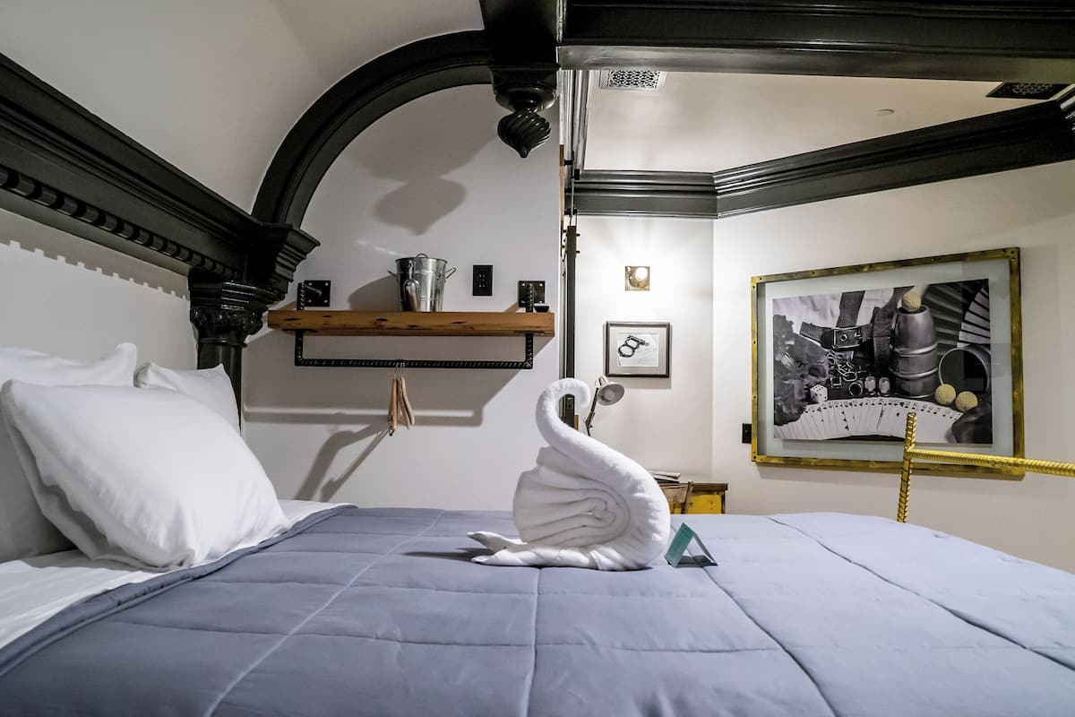 Een keurig opgemaakt bed met een handdoek in de vorm van een zwaan, een ingelijste foto aan de muur en een houten plank met een waterkoker.