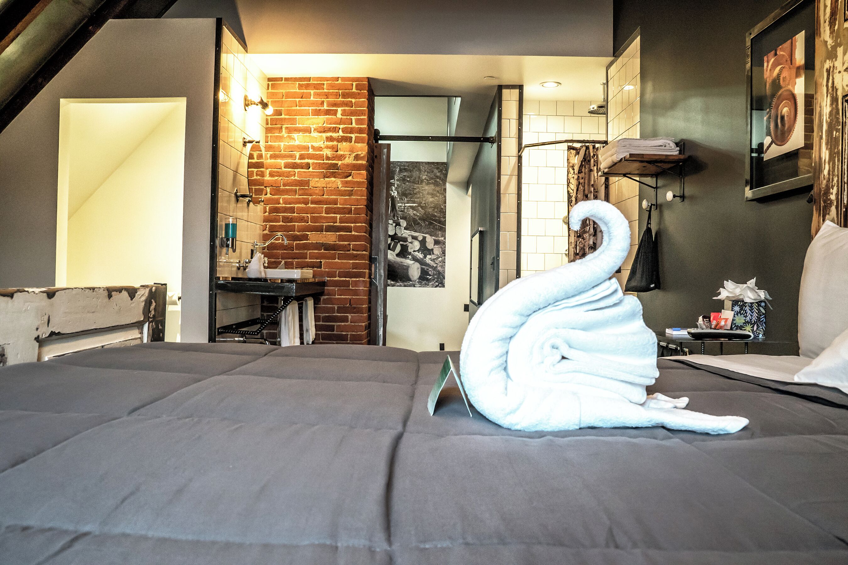 A hotel room with a neatly made bed, a brick wall, a bathroom with a mirror, and a painting on the wall.