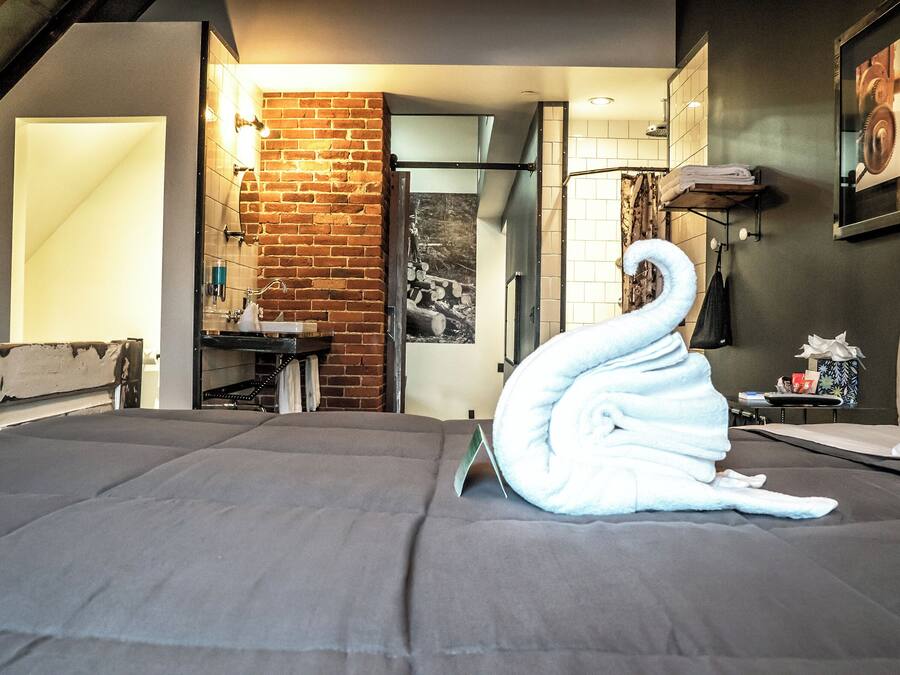 A hotel room with a neatly made bed, a brick wall, a bathroom with a mirror, and a painting on the wall.