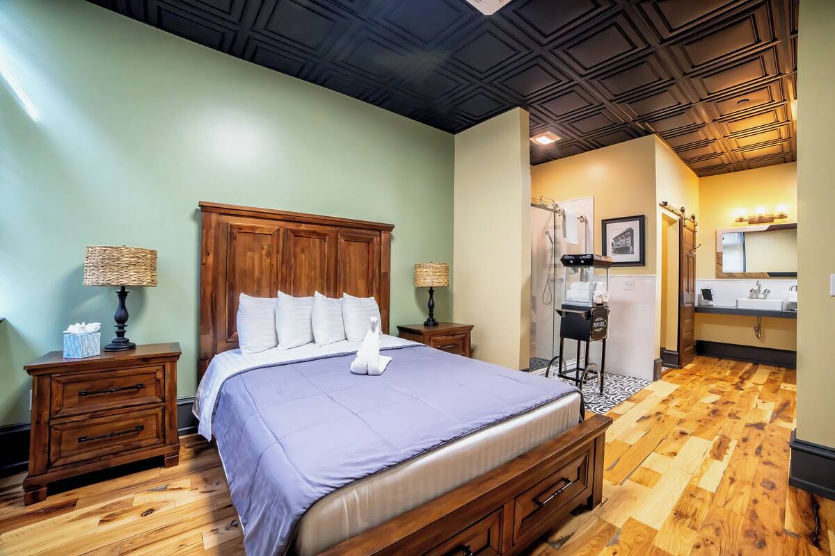 Een hotelkamer met een houten bed, een nachtkastje met een lamp, een badkamer met een wastafel en spiegel, en een houten vloer.