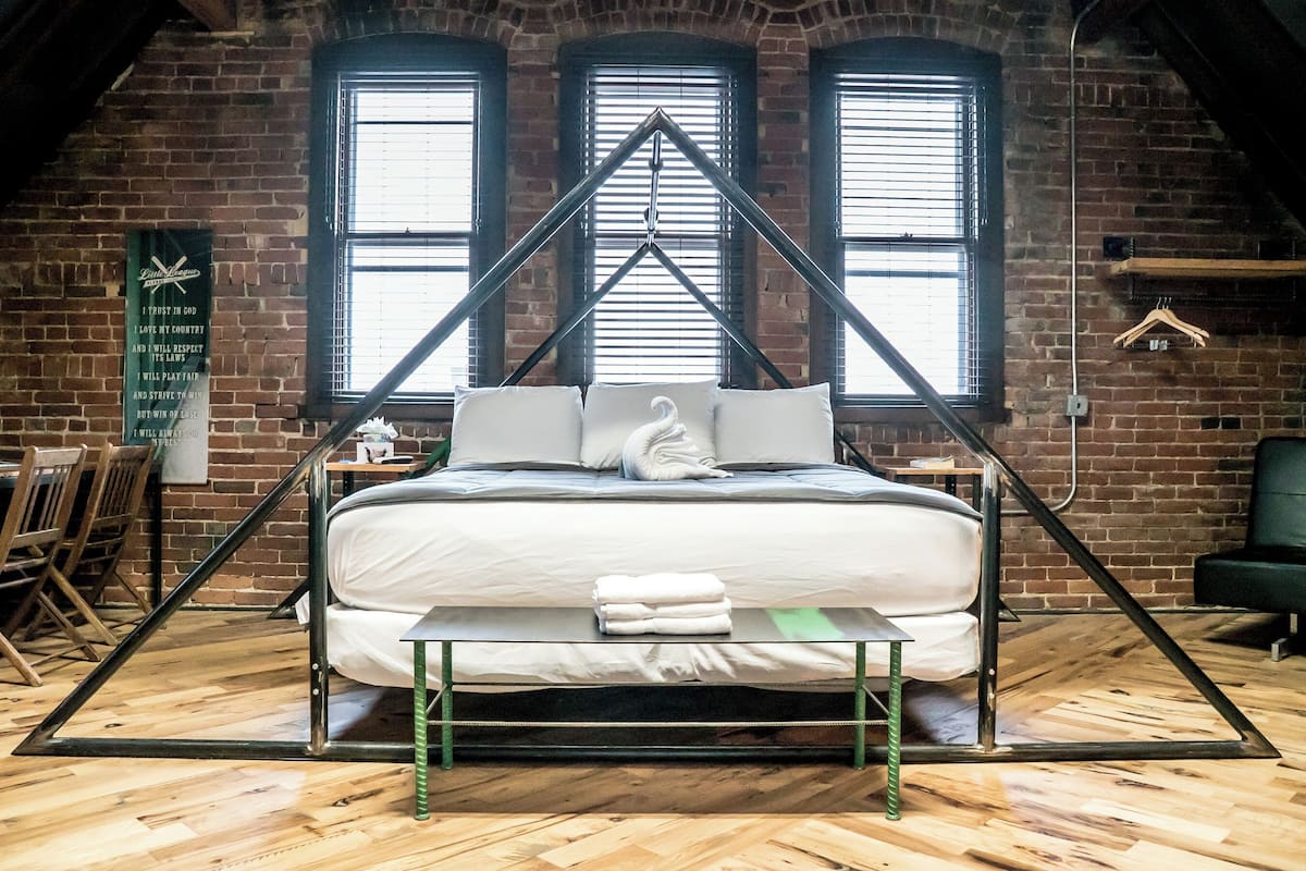 Een bed met een geometrisch frame in een kamer met bakstenen muren en grote ramen.