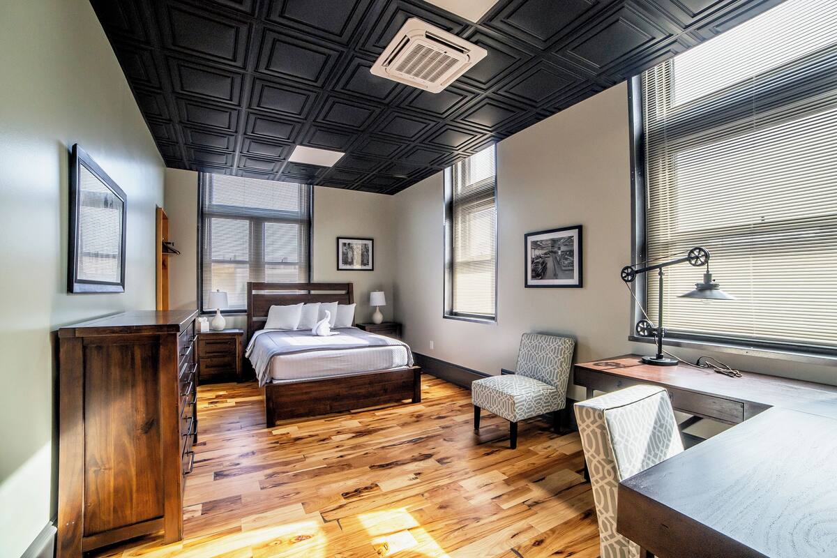 Een slaapkamer met een houten vloer, een bed met witte lakens, een bureau met lamp en een stoel.