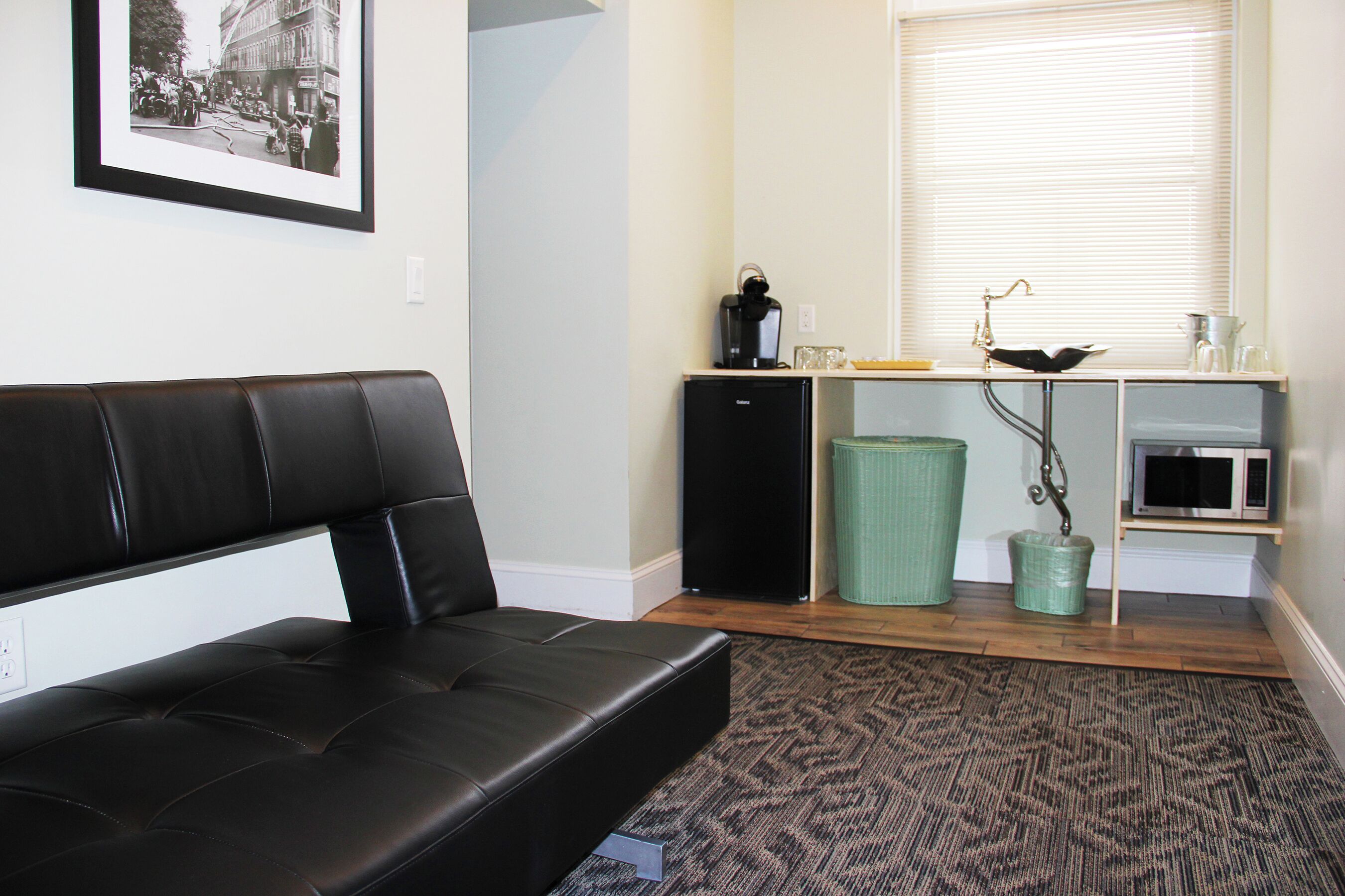 A compact living space with a black leather sofa, a small black refrigerator, a microwave, a sink, and a framed picture on the wall.
