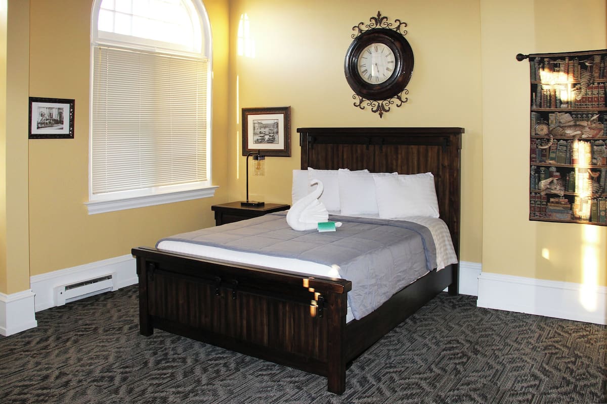 Een keurig opgemaakt bed met een houten hoofdbord, een grote wandklok en een boekenplank vol boeken.