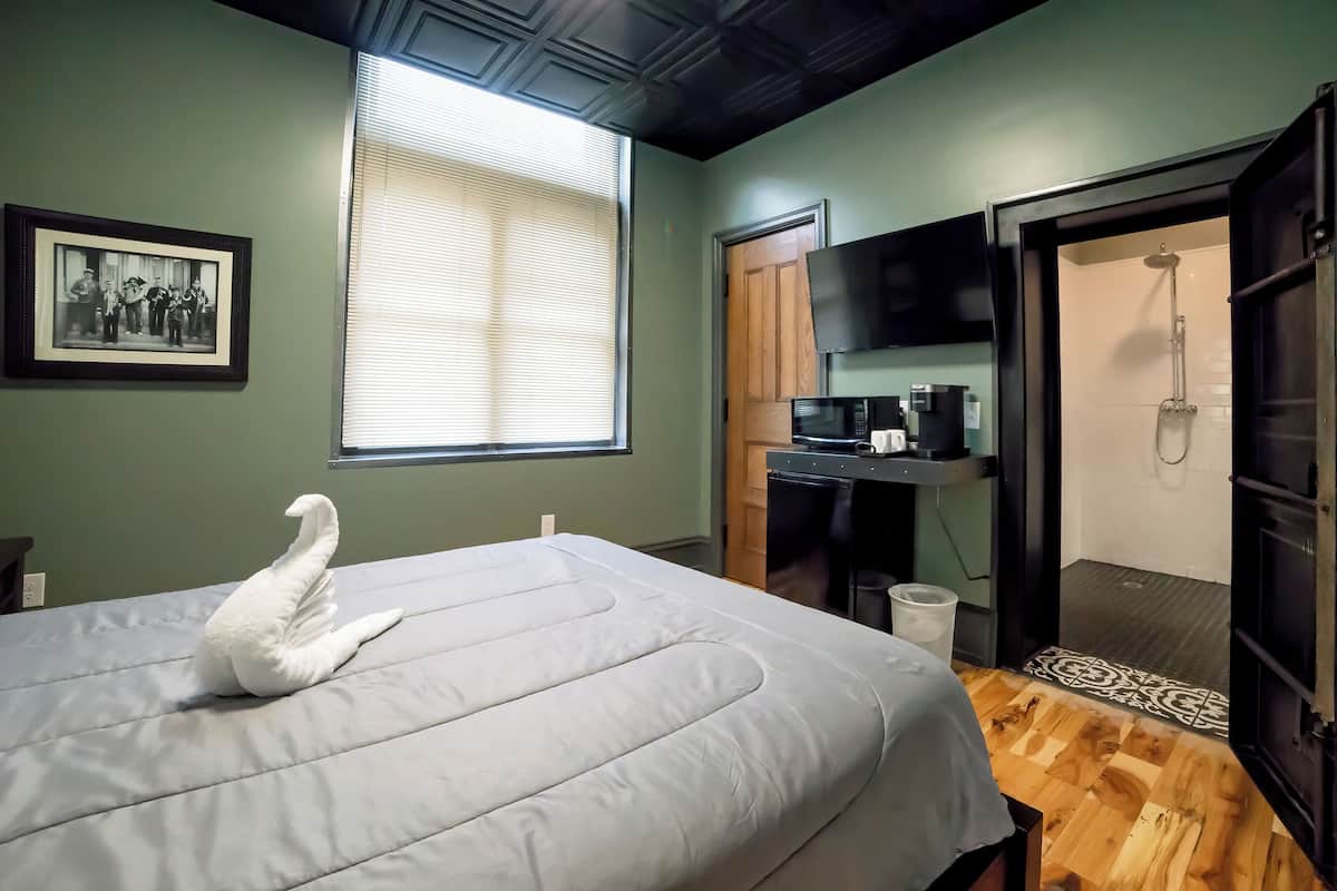 Een slaapkamer met een bed, een ingelijste foto aan de muur, een raam met zonwering, een douche en een kleine kitchenette.