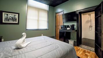 A bedroom with a bed, a framed photo on the wall, a window with blinds, a shower area, and a small kitchenette.