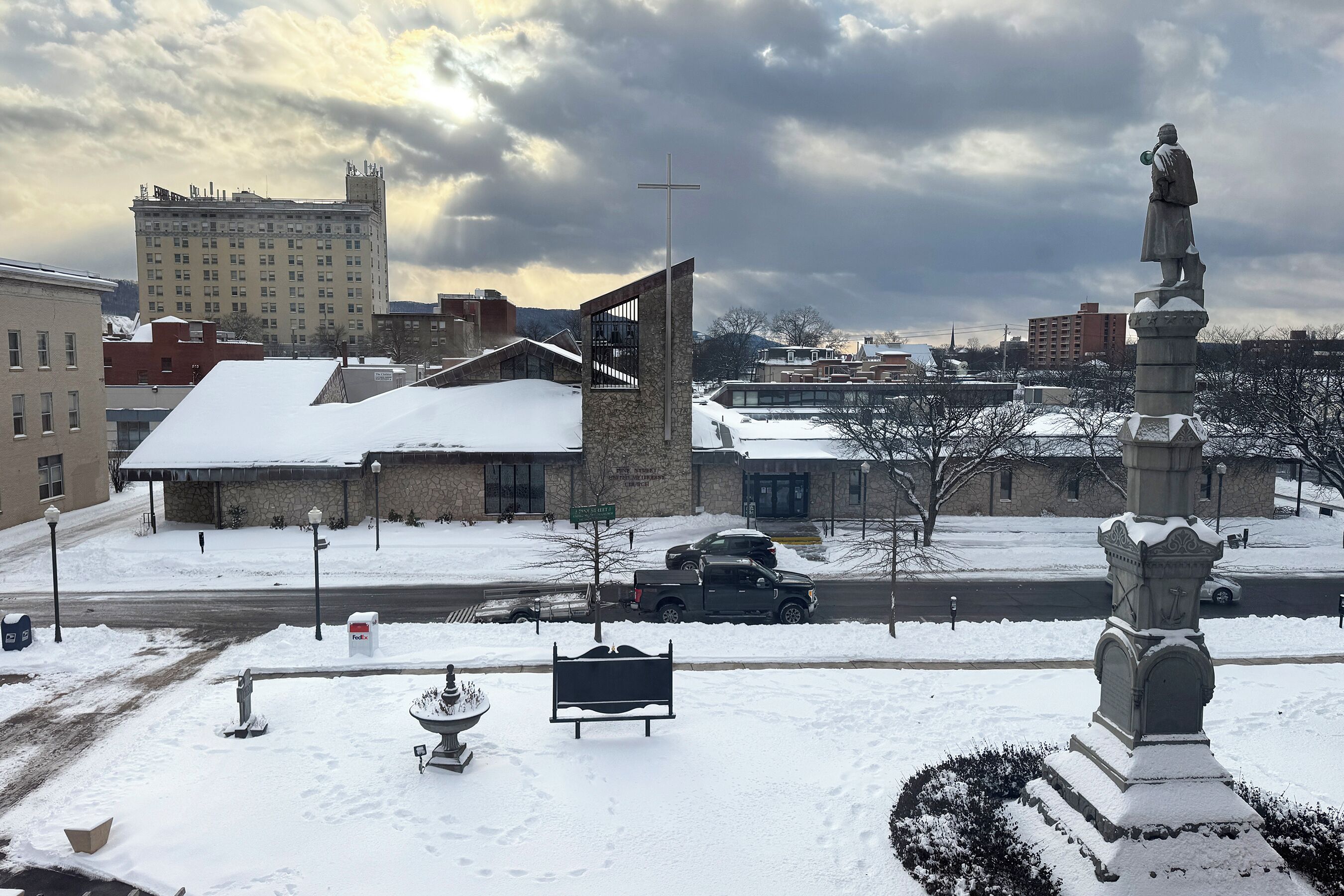 Area esterna innevata con una statua, una panchina e un edificio sullo sfondo.