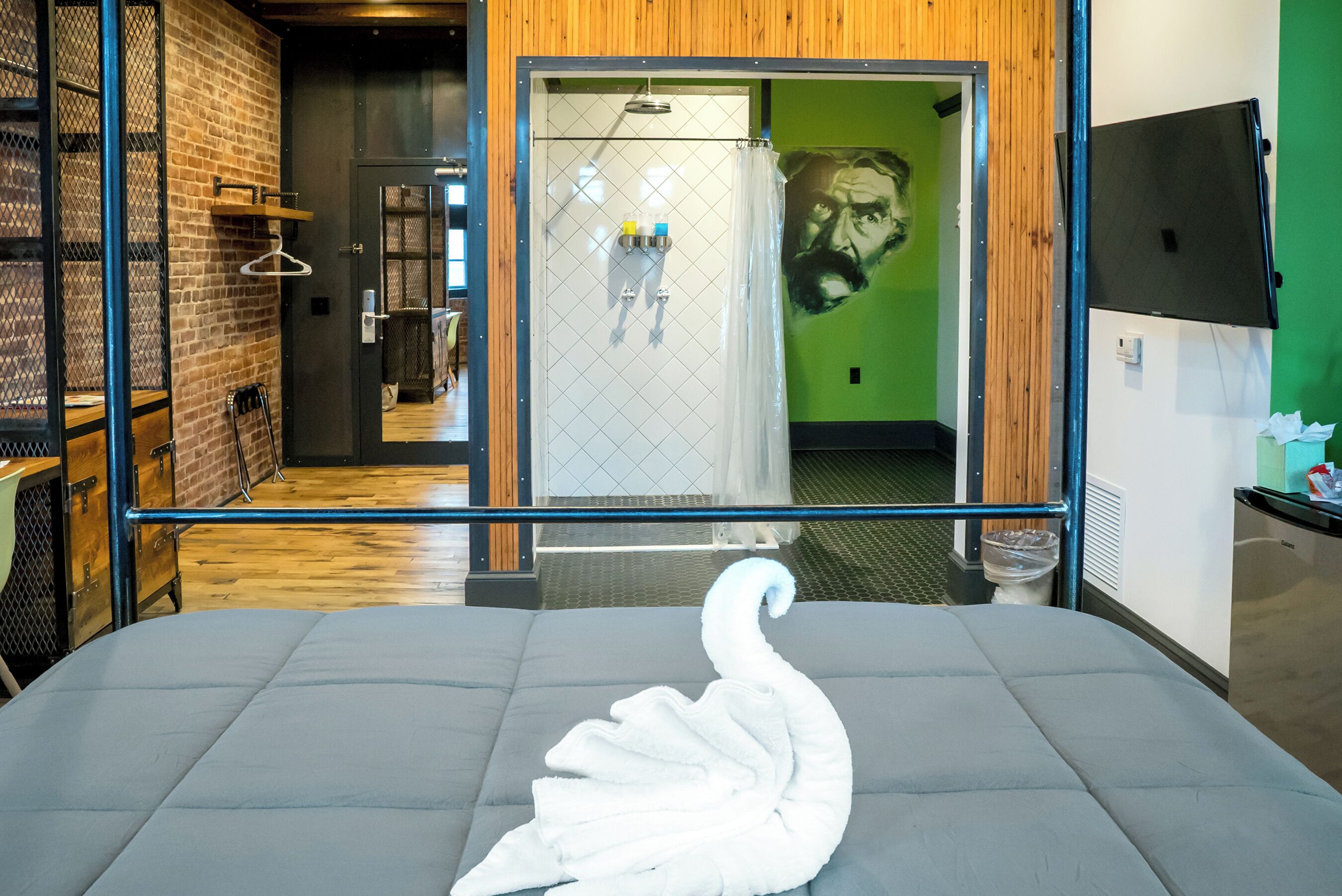 Une chambre d’hôtel avec un lit gris, une serviette blanche en forme de cygne, un espace douche et un mur vert orné d’un portrait.