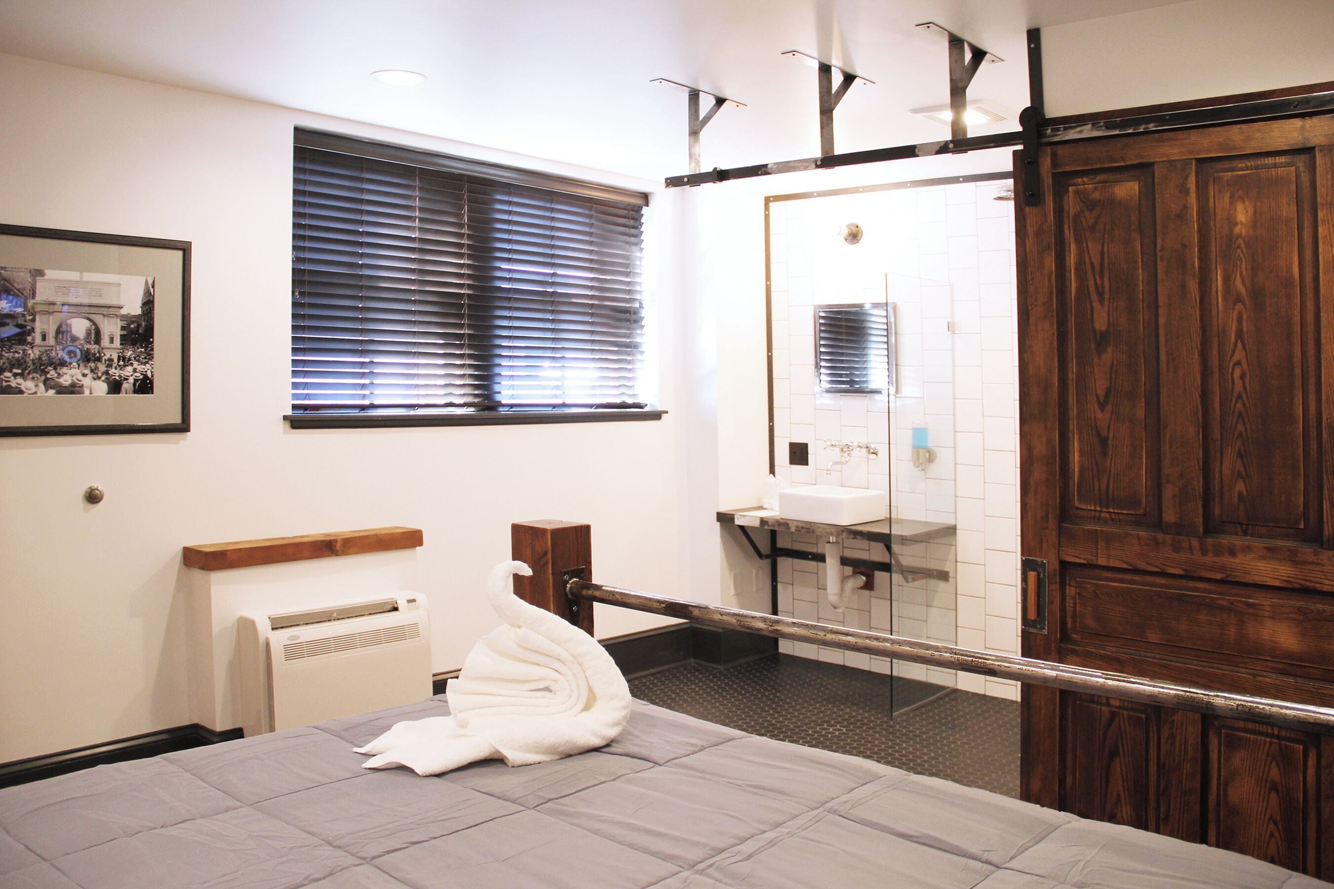 A hotel room with a bed, a window with blinds, a bathroom with a sink and mirror, and a wooden door.