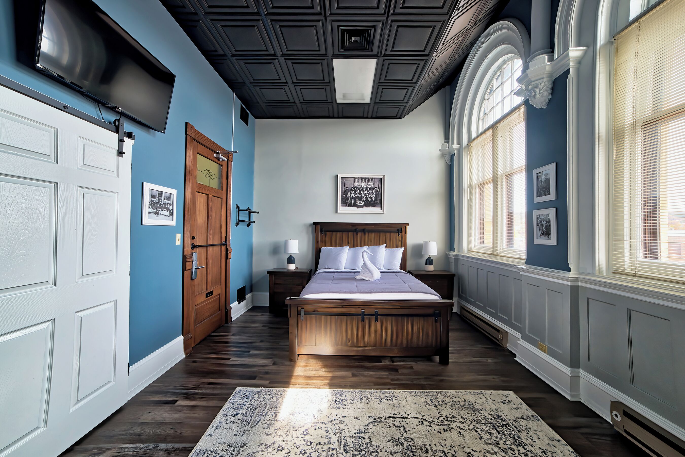 A bedroom with a wooden bed, a large window with blinds, a ceiling with a grid pattern, and a wall-mounted TV.