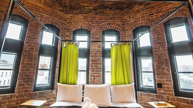 Een kamer met een hoge plafond, zichtbare bakstenen muren en grote ramen met groene gordijnen.