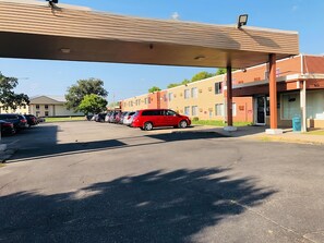 A parking lot with a row of cars, a red car parked under a canopy, and a building with multiple windows and a door. - Econo Lodge Waite Park - St. Cloud (Waite Park)
