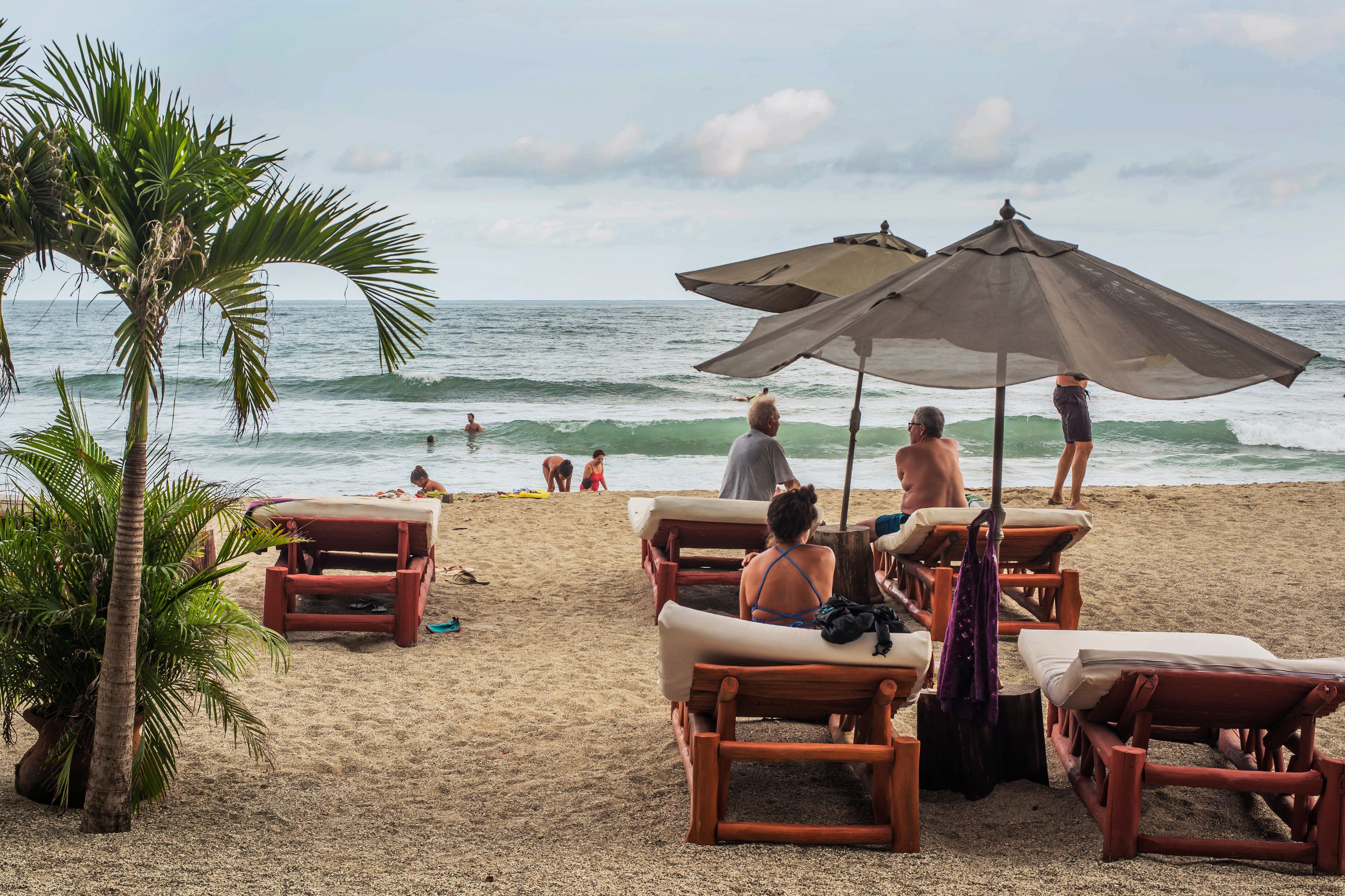A beach with wooden lounge chairs and umbrellas, palm trees, and the ocean in the background.