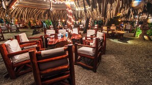 An outdoor dining area with wooden chairs and tables, surrounded by thatched roof and string lights. - Hotel Locanda Samara Beach (Sámara)