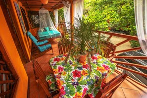 Un balcón con una mesa cubierta por un mantel floral, rodeado de plantas en macetas y una silla azul.