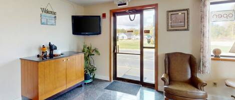 Zona de recepción con un escritorio de madera, un televisor, una planta en maceta, un sillón de cuero y una puerta que da al exterior.