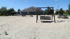 Una zona de playa arenosa con un conjunto de columpios de madera, mesas de picnic y un edificio al fondo.